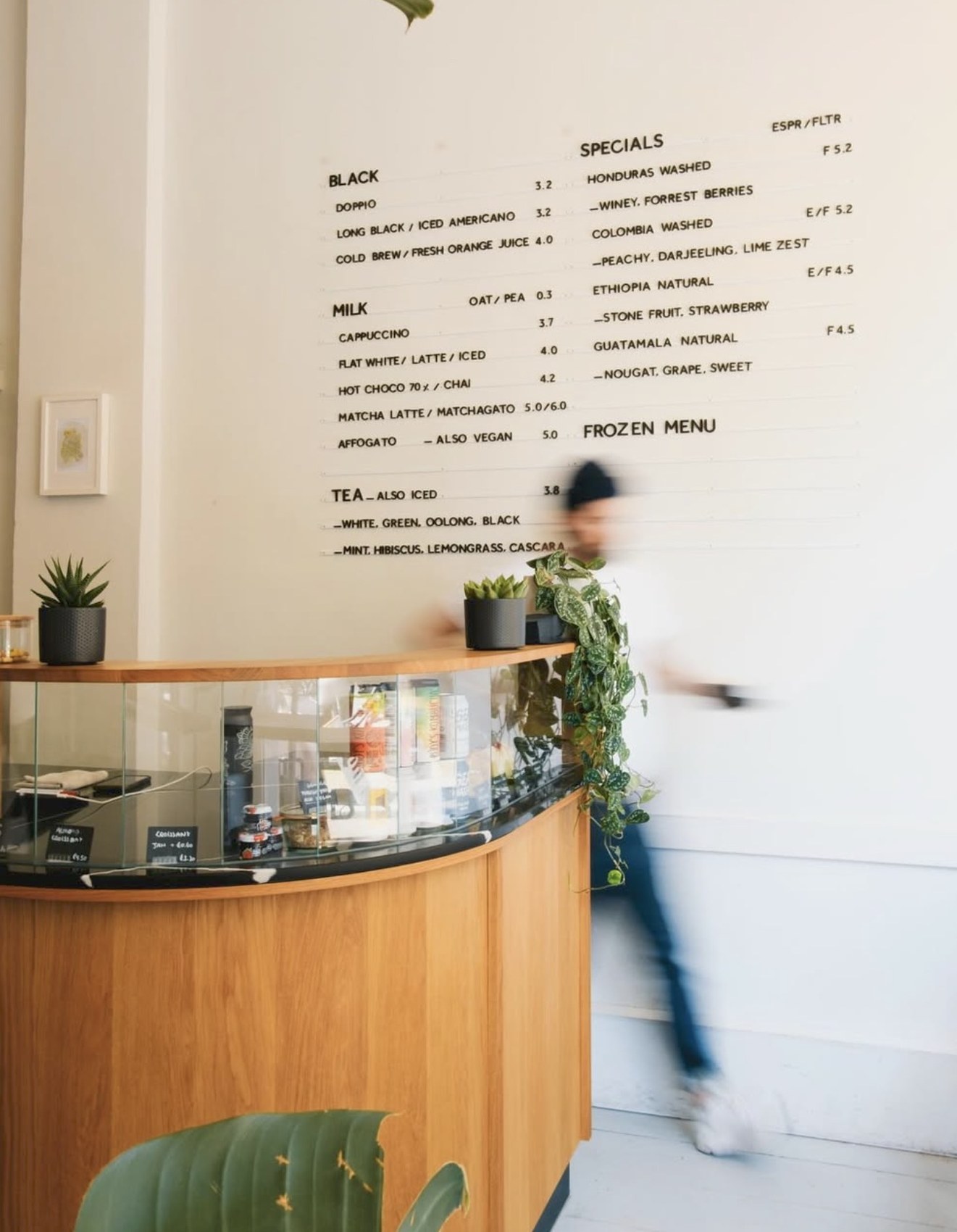 The hand-lettered menu wall above the curved wooden counter, a barista in motion carrying something past in the background